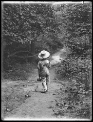 William Gray Hassler i stråhatt og kjeledress, bærende fiskestang og kurv, gående nedover en sti i skogen alene, 20. august 1911